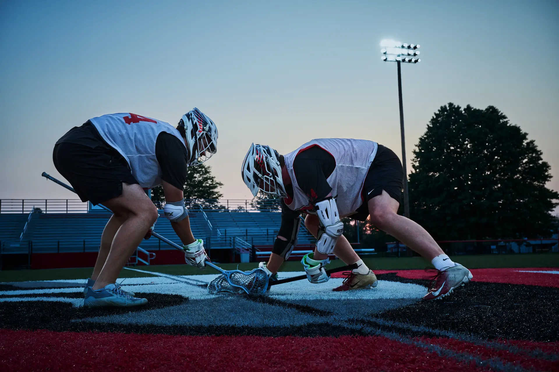 Two men wearing Lacrosse gear face off on a field at dusk