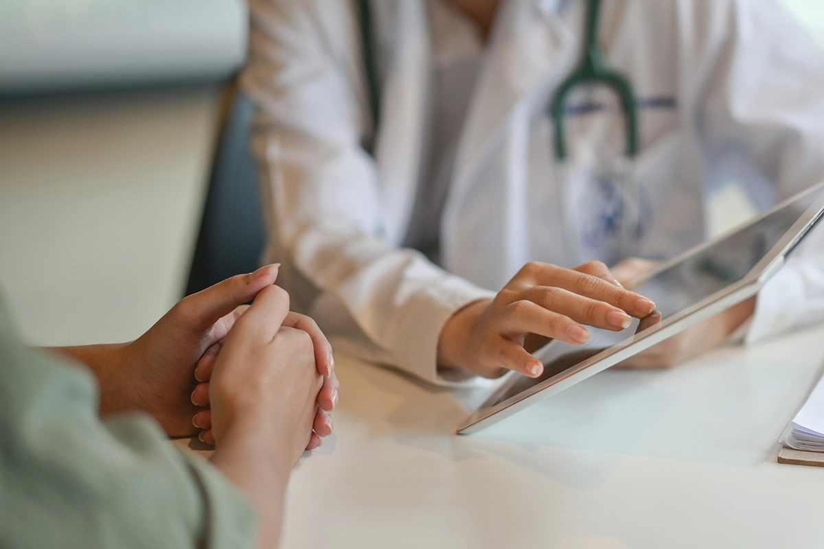 Clasped hands of a woman next to hands of a a female healthcare provider holding a tablet