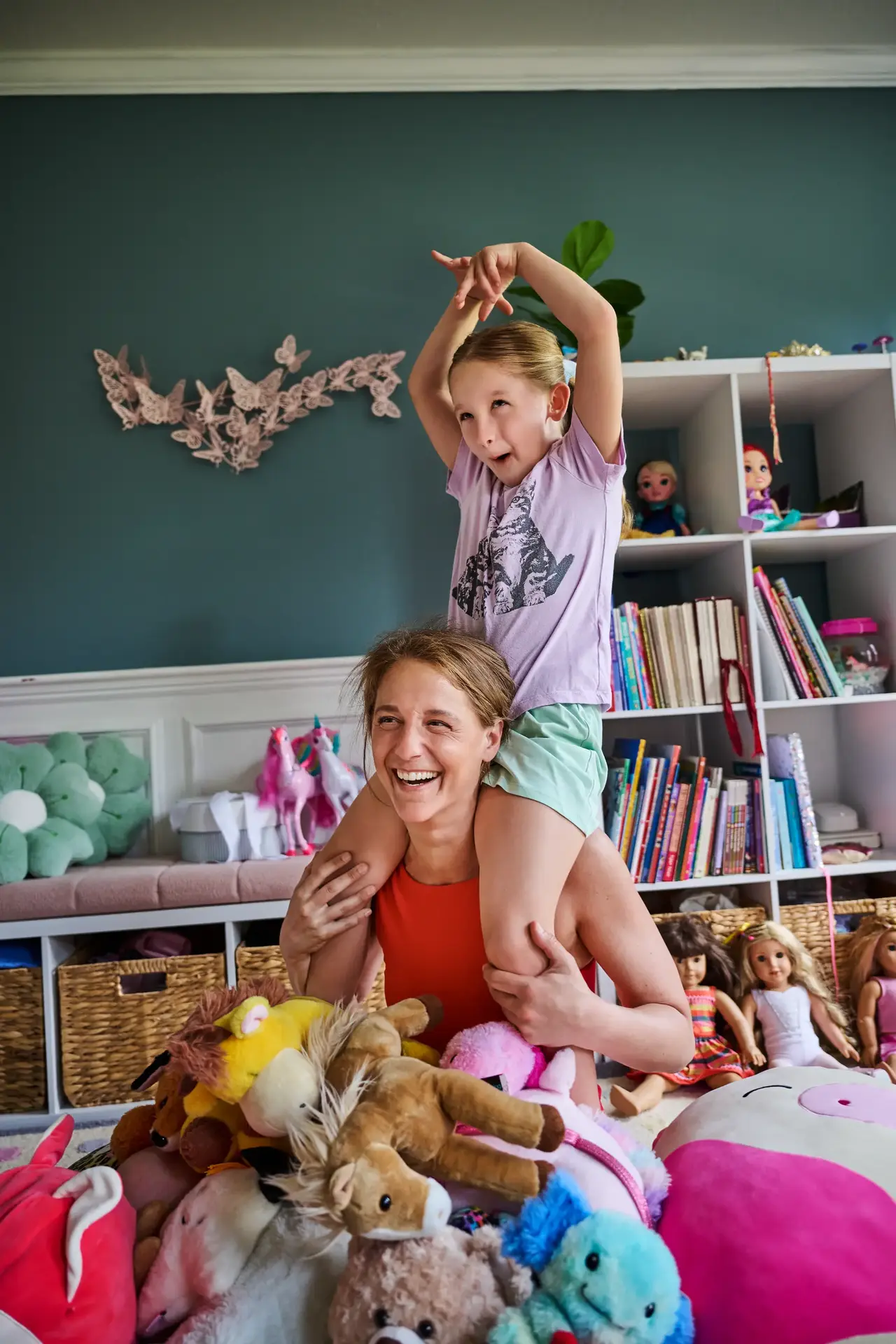 A woman sitting indoors with a child on her shoulders, and stuffed animals all around