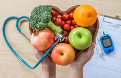 A heart shaped bowl held in a pair of hands, filled with colorful fruits and vegetables. On the table is a blood glucose meter, a clipboard, and a form.