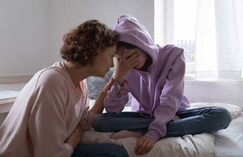 Image of a young girl sitting on her bed with her head in her hand, being comforted by her mother.