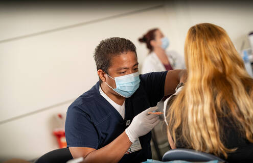 Male nurse wearing a mask administers a flu shot to a female patient with her back turned toward the camera