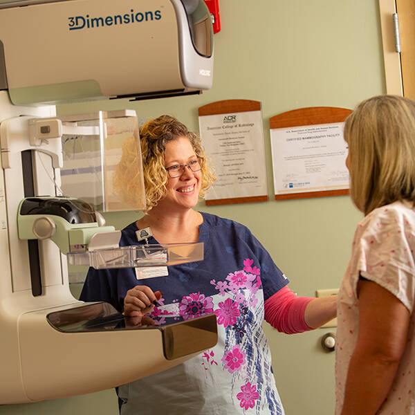 A provider stands with a patient next to a 3D mammogram machine