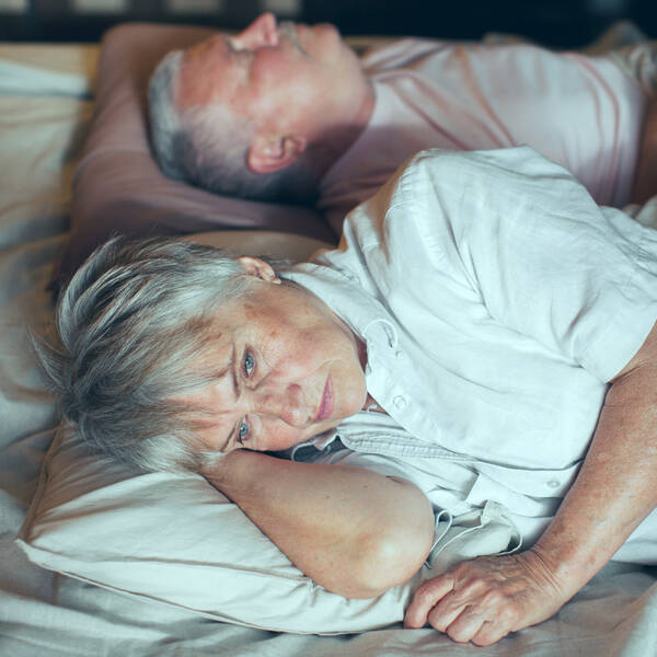 an older couple in bed, the woman lies awake while the man sleeps