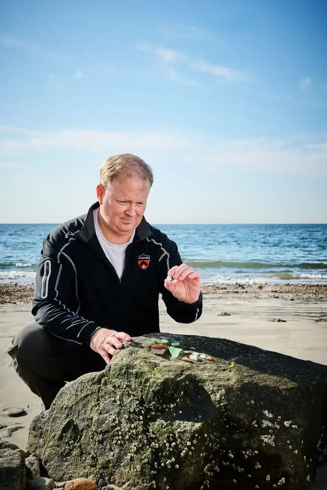 Jeff Clark crouching in front of a rock at the ocean, examining sea glass