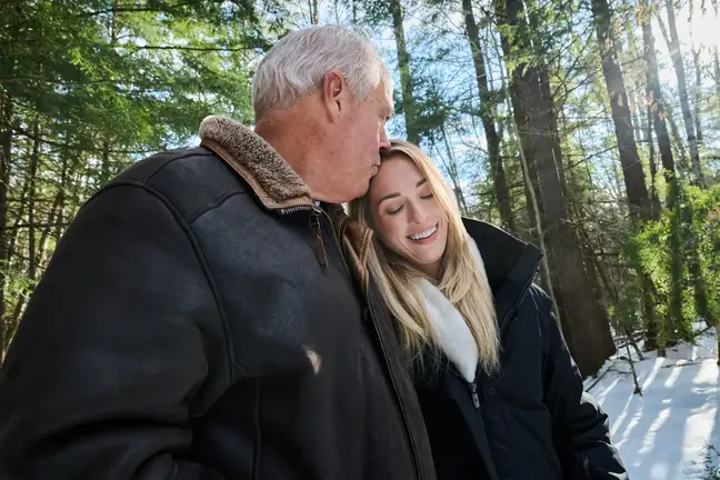 An older man kisses the forehead of a younger woman outdoors on a sunny but snow-covered Winter's day