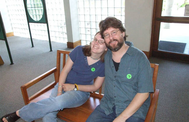 A woman and a man are sitting on a wooden bench indoors, smiling at the camera. The woman is leaning her head on the man's shoulder, and both are wearing casual clothing with green stickers on their shirts. The background features glass block windows and a door with a window.