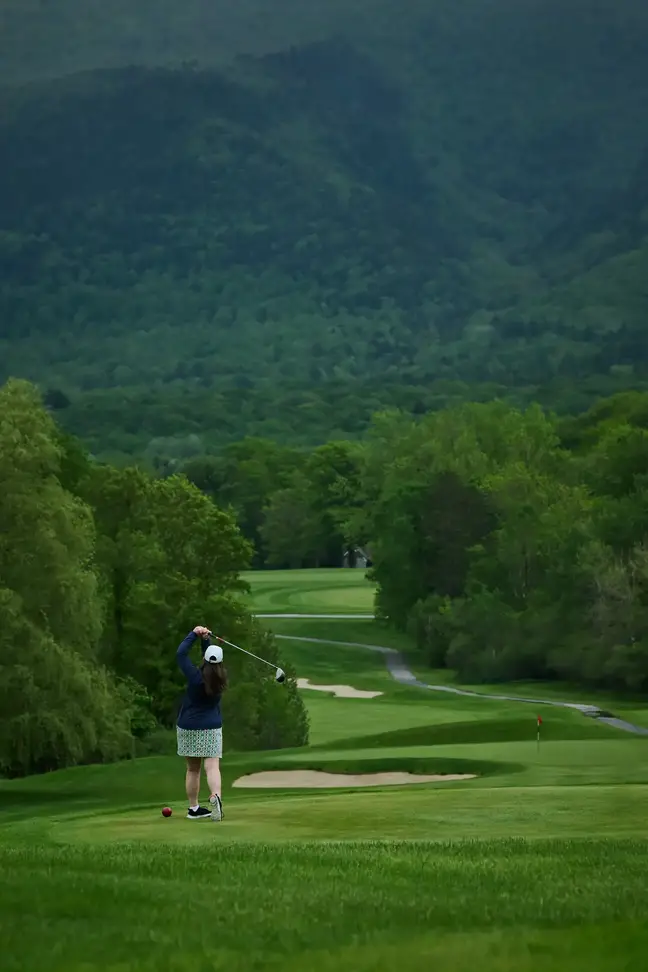 A woman hits a golf ball on a green with lush mountains in the background