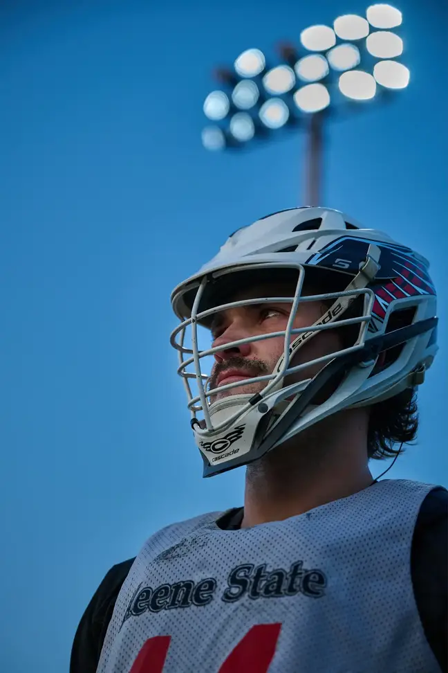 A man wearing a Lacrosse helmet, in front of field lights at dusk, looks off into distance