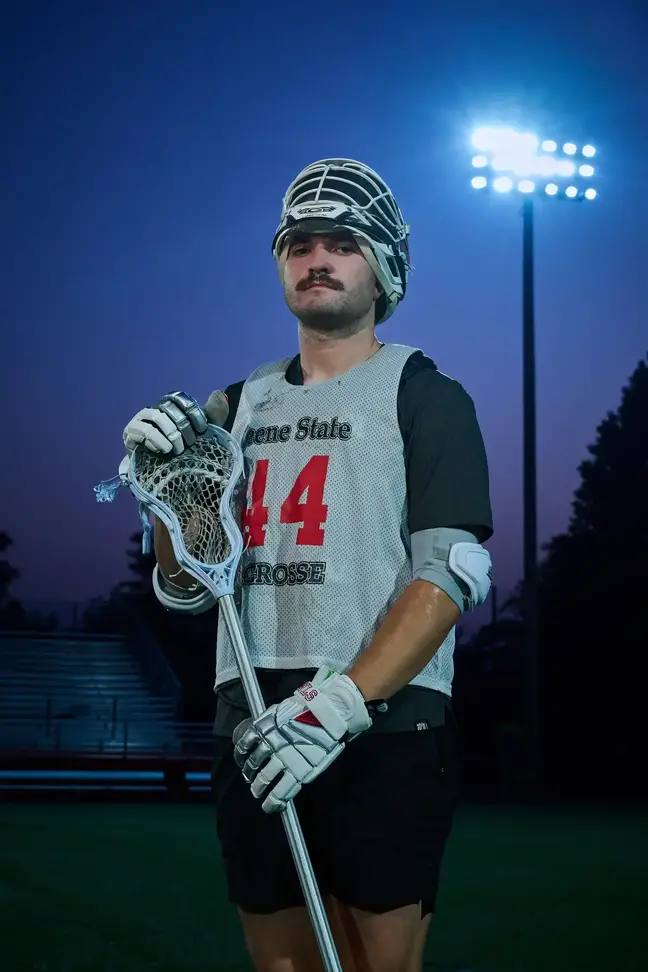 Man standing in full Lacrosse gear at dusk, with field lighting behind him