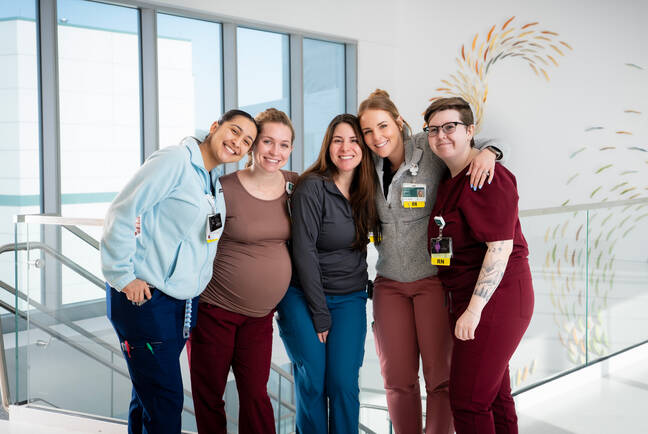 A group of five nurses smiling and standing together in a modern, bright hospital corridor. They are wearing scrubs and identification badges, showing camaraderie and teamwork.