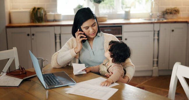 A woman is sitting at a kitchen table, talking on the phone while holding a small child who is reaching towards some papers on the table. There is a laptop, a mug, and some notebooks on the table. The background shows kitchen cabinets and a window with sunlight streaming in.