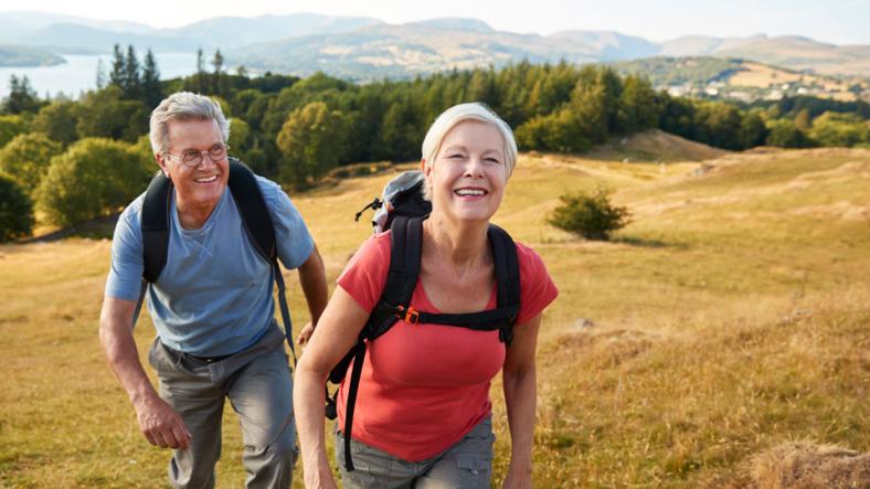 Woman and man wearing backpacks, walking in a field with mountains in the background